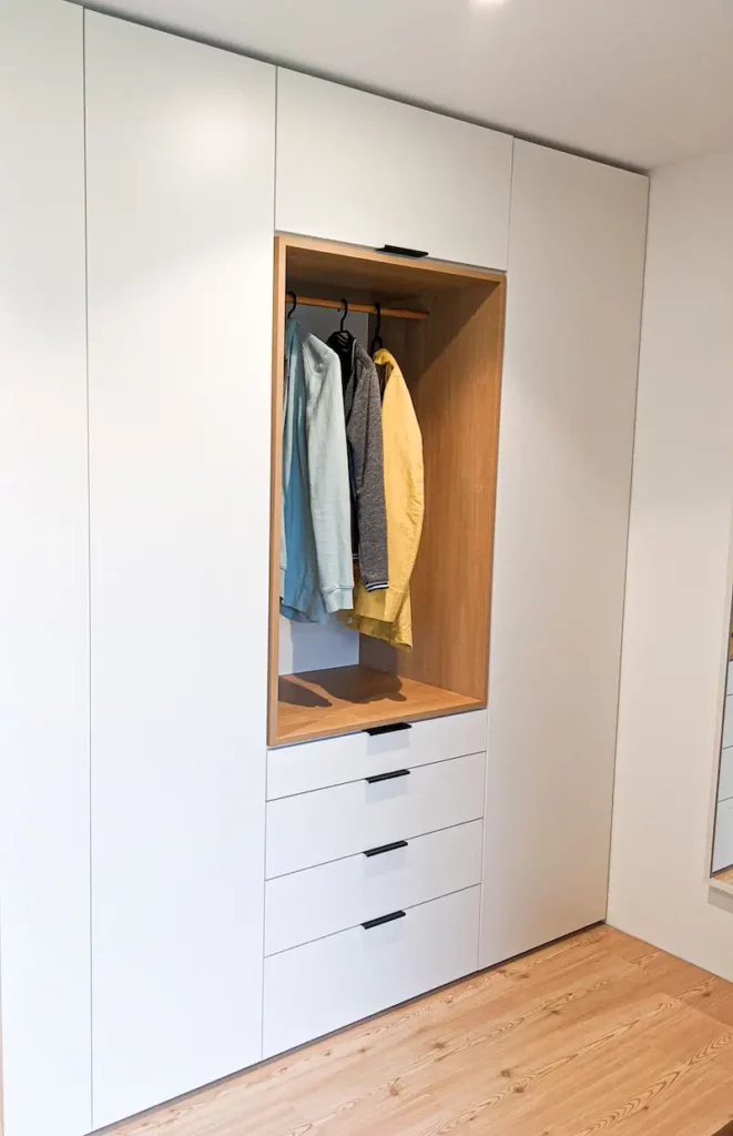 Garderobenschrank in Weiss mit Einschub aus Holz und Kleidung.
